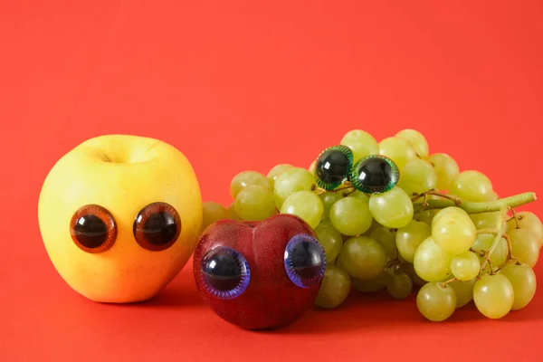 plum, apple and bunch of grapes with puppet eyes, prank concept, red background, april 1 prank, cute fruit faces