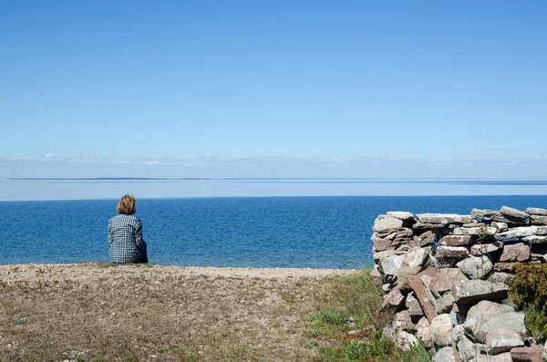 Woman sitting by the coast looking at clear blue sky and water — Stock Photo, Image