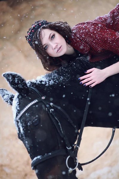 Bella e bella ragazza dell'aspetto europeo bruna con cavallo marrone nella natura invernale con accessori. Moda e bellezza. Animale e naturale . Immagine Stock