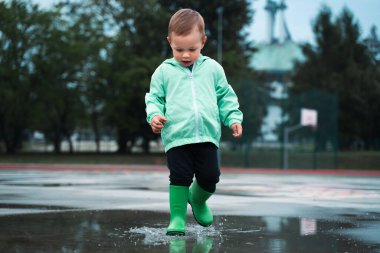 Boy playing outside summer rain time. Children is wearing green raincoat and Rubber boots. Kid having fun at rainy day. Childchood, autumn concept.