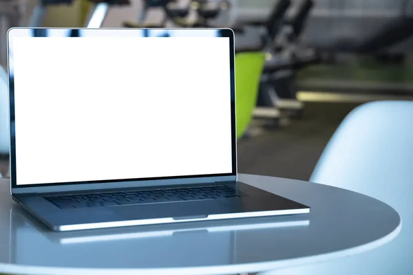 Laptop with white screen in fitness center in shopping mall. Empty copy space, blank screen mockup. Soft focus laptop with interor background. Healthy, gym and yoga concept.