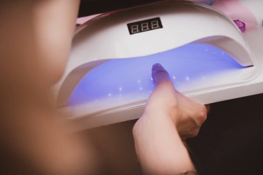 Manicurist master is covering painting client's nails top coat shellac, hands closeup. One hand drying in UV lamp. Professional manicure with gel polish. Nail art, hands care, beauty industry concept.