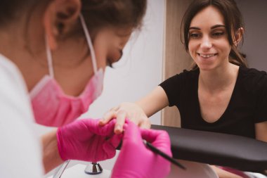 Manicurist master is covering painting client's nails top coat shellac, hands closeup. One hand drying in UV lamp. Professional manicure with gel polish. Nail art, hands care, beauty industry concept.