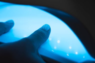 Manicurist master is covering painting client's nails top coat shellac, hands closeup. One hand drying in UV lamp. Professional manicure with gel polish. Nail art, hands care, beauty industry concept.