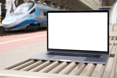 Laptop with white screen in railway station. Empty copy space, blank screen mockup. Soft focus laptop with train background. Travel, study and work outside office concept.