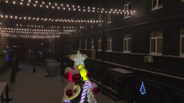 Árvore de Natal decorada na rua nevada durante a queda de neve. Decoração de árvore de Natal com brinquedos, caramelos de cana de doces, bolas e homem de gengibre. Iluminados brinquedos de árvore de ano novo — Vídeo de Stock