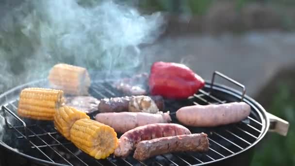 Smažené klobásy s kukuřicí a červeným pepřem na kulatém černém grilu, zakryté víkem, s kouřem a krajinou na venkově — Stock video