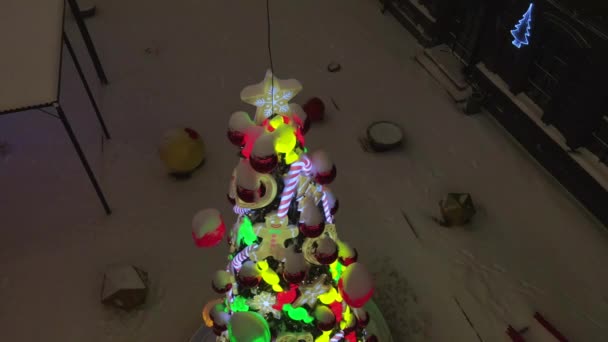 Kar yağışı sırasında karlı caddede süslenmiş Noel ağacı. Oyuncaklarla Noel ağacı süslemesi, şeker kamışlı karamel, top ve zencefilli kurabiye adam. Aydınlatılmış yeni yıl ağacı oyuncakları. 4k drone videosu — Stok video