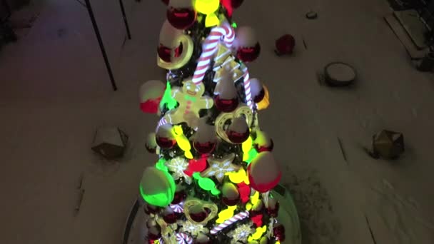 Albero di Natale decorato sulla strada innevata durante le nevicate. Decorazione dell'albero di Natale con giocattoli, caramelle di canna da zucchero, palline e uomo pan di zenzero. Giocattoli di albero illuminati di anno nuovo. 4k drone video — Video Stock