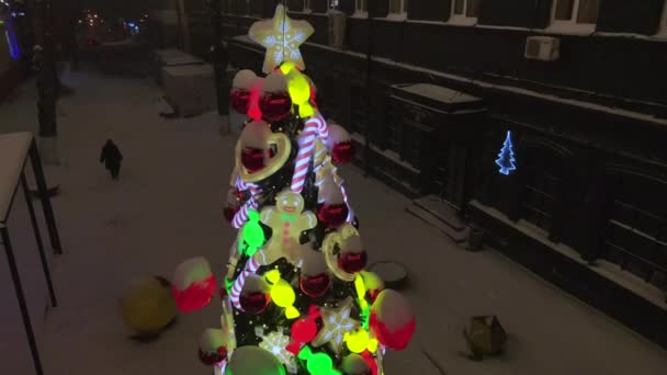 Geschmückter Weihnachtsbaum auf verschneiter Straße bei Schneefall. Christbaumschmuck mit Spielzeug, Zuckerrohrkaramellen, Kugeln und Lebkuchenmann. Beleuchtetes Silvesterspielzeug. 4k Drohnenvideo — Stockvideo