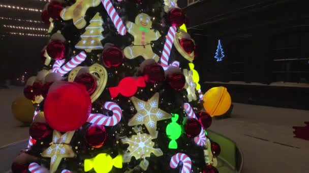Ozdobiona choinka na śnieżnej ulicy podczas opadów śniegu. Choinka ozdobiona zabawkami, karmelkami z trzciny cukrowej, kulkami i piernikiem. Oświetlone noworoczne zabawki. 4k wideo drona — Wideo stockowe