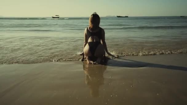 Νεαρή λεπτή όμορφη κοκκινομάλλα κοπέλα με λευκό μαγιό κάνει ηλιοθεραπεία το ηλιοβασίλεμα καθισμένη στην παραλία — Αρχείο Βίντεο