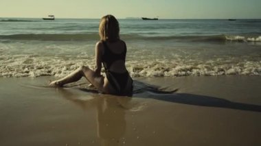 Sıska, güzel, kızıl saçlı, beyaz mayo giyen kız gün batımında güneşleniyor plajda oturuyor.