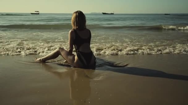 Joven delgado hermosa pelirroja chica en un traje de baño blanco toma el sol al atardecer sentado en la playa — Vídeo de stock