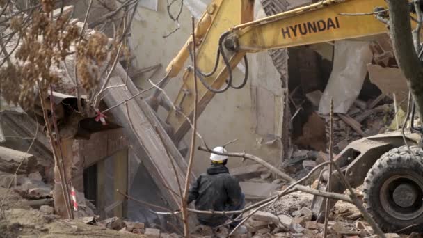 Engenheiro com capacete de proteção olha para a demolição, destruição de edifício antigo, escavadeira quebra a casa do vintage. Desconstrução do objeto vivo. Renovação urbana 4k Vídeo em câmera lenta de alta qualidade — Vídeo de Stock