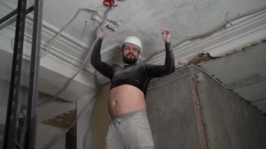 Koruyucu kasklı ve siyah dar elbiseli şişman sakallı yetişkin adam inşaat beyazlatıcısının yüksek tavanını kaldırıyor. Tamiratı kendi el konseptinle yap. 4k ağır çekim görüntüleri 