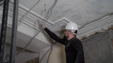Koruyucu kasklı ve siyah dar elbiseli şişman sakallı yetişkin adam inşaat beyazlatıcısının yüksek tavanını kaldırıyor. Tamiratı kendi el konseptinle yap. 4k ağır çekim görüntüleri 