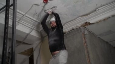 Koruyucu kasklı ve siyah dar elbiseli şişman sakallı yetişkin adam inşaat beyazlatıcısının yüksek tavanını kaldırıyor. Tamiratı kendi el konseptinle yap. 4k ağır çekim görüntüleri 