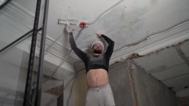 Koruyucu kasklı ve siyah dar elbiseli şişman sakallı yetişkin adam inşaat beyazlatıcısının yüksek tavanını kaldırıyor. Tamiratı kendi el konseptinle yap. 4k ağır çekim görüntüleri 