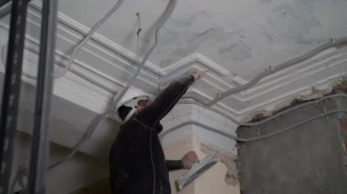 Koruyucu kasklı ve siyah dar elbiseli şişman sakallı yetişkin adam inşaat beyazlatıcısının yüksek tavanını kaldırıyor. Tamiratı kendi el konseptinle yap. 4k ağır çekim görüntüleri 
