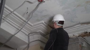 Koruyucu kasklı ve siyah dar elbiseli şişman sakallı yetişkin adam inşaat beyazlatıcısının yüksek tavanını kaldırıyor. Tamiratı kendi el konseptinle yap. 4k ağır çekim görüntüleri 