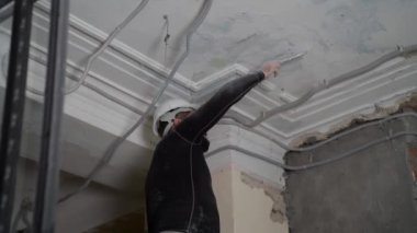 Koruyucu kasklı ve siyah dar elbiseli şişman sakallı yetişkin adam inşaat beyazlatıcısının yüksek tavanını kaldırıyor. Tamiratı kendi el konseptinle yap. 4k ağır çekim görüntüleri 