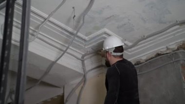 Koruyucu kasklı ve siyah dar elbiseli şişman sakallı yetişkin adam inşaat beyazlatıcısının yüksek tavanını kaldırıyor. Tamiratı kendi el konseptinle yap. 4k ağır çekim görüntüleri 