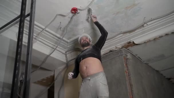 Fat bearded adult man in protective helmet and black tight fitting clothing remove construction whitewash vintage high ceiling. Overhaul repair with your own hands concept. 4k slow motion footage — Stock Video