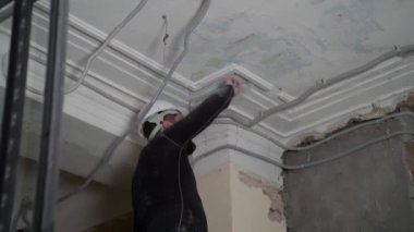 Koruyucu kasklı ve siyah dar elbiseli şişman sakallı yetişkin adam inşaat beyazlatıcısının yüksek tavanını kaldırıyor. Tamiratı kendi el konseptinle yap. 4k ağır çekim görüntüleri 