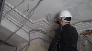 Koruyucu kasklı ve siyah dar elbiseli şişman sakallı yetişkin adam inşaat beyazlatıcısının yüksek tavanını kaldırıyor. Tamiratı kendi el konseptinle yap. 4k ağır çekim görüntüleri 