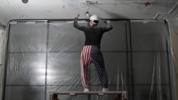 Жирний бородатий дорослий чоловік в захисному шоломі і прапор США друк на штанях видаляють будівельні білизна вінтажний високий стелю. Ремонт здійснюється за допомогою концепції власних рук. 4k повільних кадрів руху — стокове відео