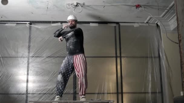 Een gelukkige Amerikaanse bouwer met een hoed op danst. Grappige werkbewegingen op de achtergrond van een bouwplaats. slow motion. — Stockvideo