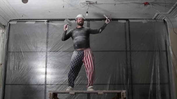 保護ヘルメットとパンツの上の米国の旗の印刷で脂肪ひげそり大人の男は、建設のホワイトウォッシュヴィンテージ高天井を削除します。あなた自身の手の概念でオーバーホール修理。4kスローモーション映像 — ストック動画