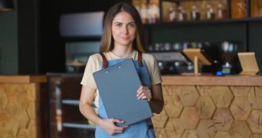 Elinde tabletle kafeteryadaki kameraya bakan önlüklü, gülümseyen garson portresi.