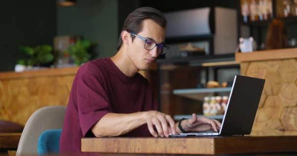 Guapo joven escribiendo en el teclado del ordenador portátil y el uso de un teléfono inteligente en la cafetería. El hombre trabaja en una computadora portátil y luego toma el teléfono móvil. Movimiento lento — Vídeos de Stock