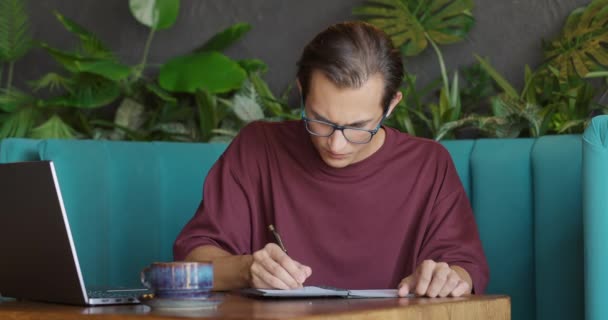 Schöner junger Mann mit Brille schreibt und zerknüllt falsch geschriebenes Papier im Café. Zeitlupe — Stockvideo