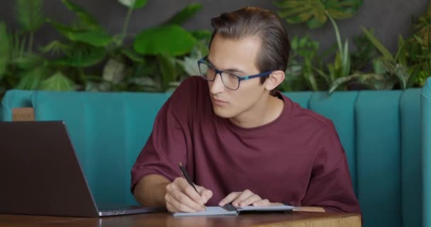Schöner junger Mann mit Brille sitzt mit Laptop an einem Tisch im Café und schreibt in Notizblock. Ferngesteuertes Bildungskonzept. Studentisches Studium. Freiberufler arbeiten. Zeitlupe — Stockvideo