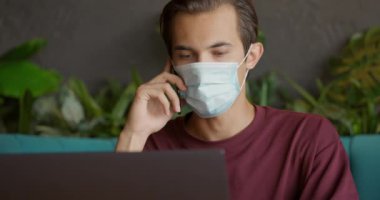Caddedeki kafede dizüstü bilgisayarla çalışan yakışıklı bir adam ve Covid-19 salgını sırasında maske takarak cep telefonuyla konuşan. İnsanlar ve iletişim konsepti.