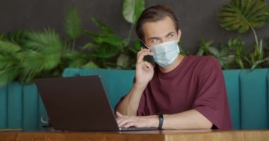 Caddedeki kafede dizüstü bilgisayarla çalışan yakışıklı bir adam ve Covid-19 salgını sırasında maske takarak cep telefonuyla konuşan. İnsanlar ve iletişim konsepti.