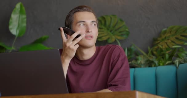 Stilig ung man i glasögon sitter vid ett bord på ett café och pratar i en röstchatt via smartphone. Killen skriver röstbrevlåda till sociala nätverk. Framifrån. Långsamma rörelser — Stockvideo