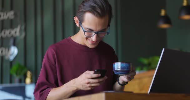 Fiatal férfi csevegés az okostelefonon és gépelés a laptopon, miközben kávészünet a kis kávézóban — Stock videók