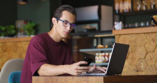 Молодий чоловік працює на ноутбуці і перевіряє смартфон на повідомлення під час перерви в кав'ярні — стокове відео