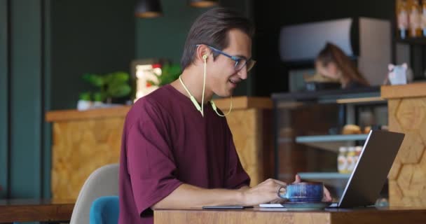 Ung stilig man som sitter på kafé med bärbar dator och gör videosamtal med webbkamera och hörlurar — Stockvideo