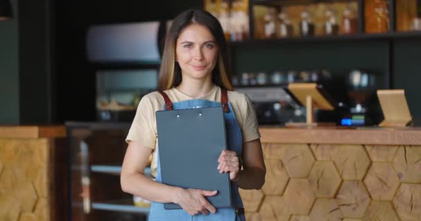 Portret van een mooie glimlachende serveerster die in een gezellig café staat en het menu vasthoudt — Stockvideo