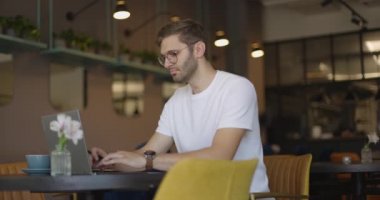Gündüz kafede oturan ve uzaktan kumanda için bilgisayarı kullanan adamın yan görüntüsü. Serbest çalışan gözlüklü işadamı, tabut içinde klavyede yazı yazıyor ve kahve içiyor.