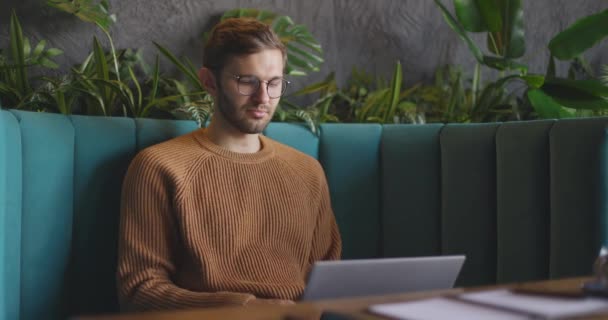Πλευρική άποψη του ανθρώπου που κάθεται στο καφέ κατά τη διάρκεια της ημέρας και χρησιμοποιώντας το φορητό υπολογιστή για την απομακρυσμένη εργασία. Όμορφος ελεύθερος επαγγελματίας επιχειρηματίας με γυαλιά που δουλεύει στο καφέ. Σπουδαστές online — Αρχείο Βίντεο