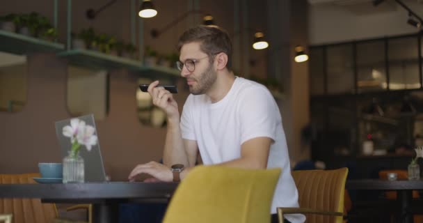ノートパソコンを使ってスマホで音声メッセージを送る喫茶店のテーブル席に座っている白いシャツを着た眼鏡姿の真剣な男。スローモーション — ストック動画