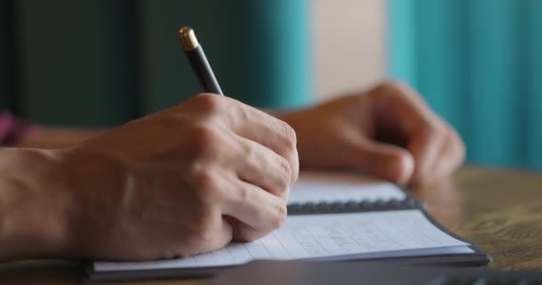 Male hands writing in paper notebook while sitting at wooden table next to the window — Stock Video
