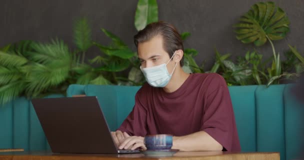 Homem usando máscara protetora trabalhando distantemente no cafe.Young cara sentado com xícara de chá e laptop — Vídeo de Stock