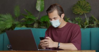 Kahve dükkanında oturan koruyucu maskeli genç bir adam. Telefonu ve laptopu kullanan yakışıklı bir öğrenci.